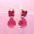 A pair of gold earrings featuring square-cut red stones suspended above larger teardrop-shaped pink gemstones rests against a soft pink backdrop.