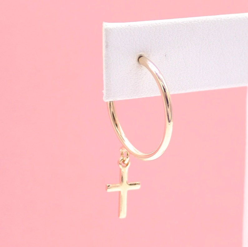 A gold hoop earring featuring a dangling cross charm is displayed against a soft pink background.