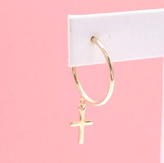 A gold hoop earring featuring a dangling cross charm is displayed against a soft pink background.