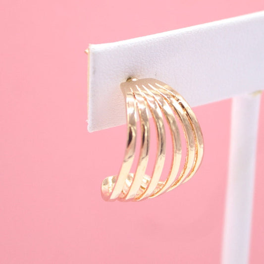 A gold multi-strand hoop earring hangs from a white display card against a soft pink background.