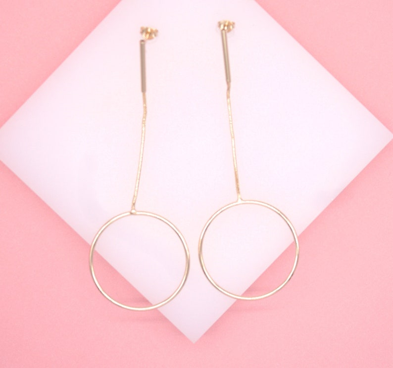 A pair of elegant gold-plated drop earrings featuring a delicate chain and circle design rests against a white display pad on a soft pink background.