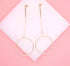 A pair of elegant gold-plated drop earrings featuring a delicate chain and circle design rests against a white display pad on a soft pink background.
