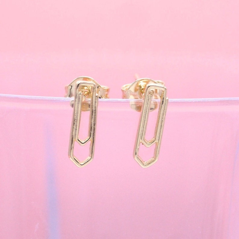 Two gold paperclip stud earrings rest against a soft pink background.