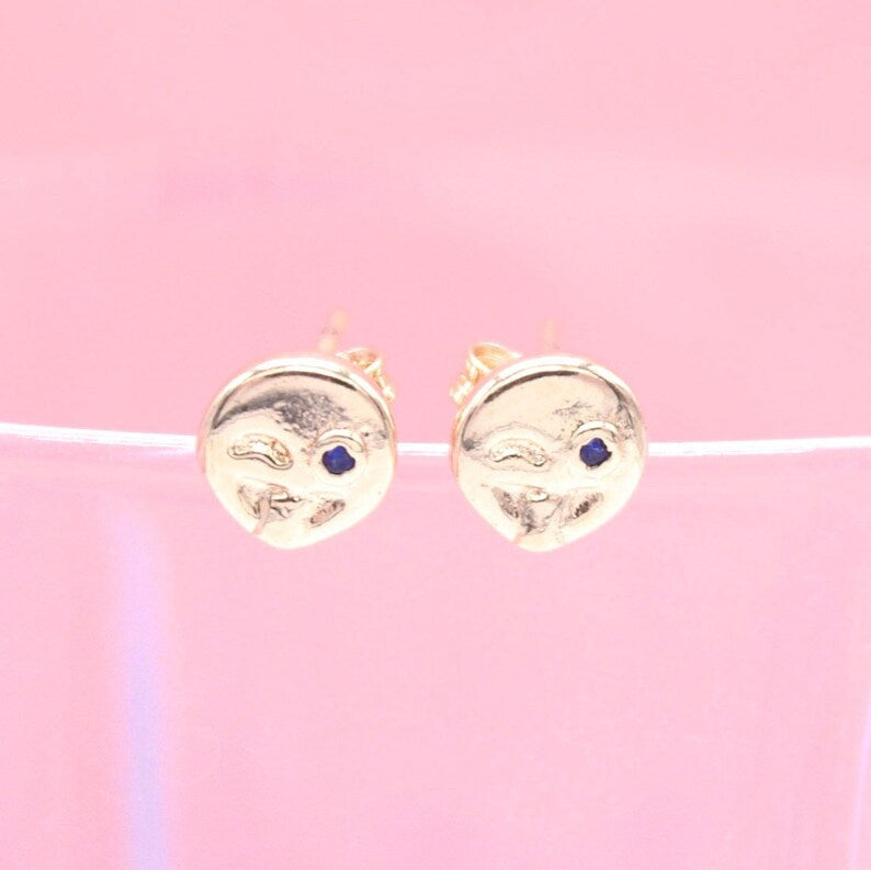 A pair of gold stud earrings featuring playful winking faces with tiny blue gemstone eyes rests against a soft pink backdrop.