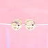 A pair of gold stud earrings featuring playful winking faces with tiny blue gemstone eyes rests against a soft pink backdrop.