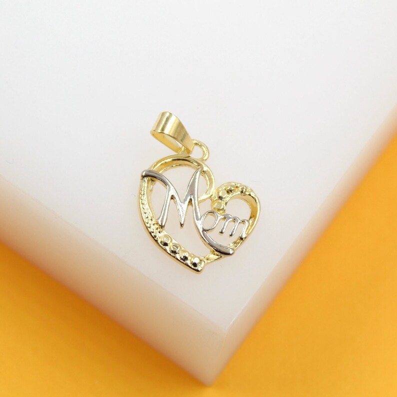 A gold and white gold heart-shaped Mom pendant rests on a white display surface.