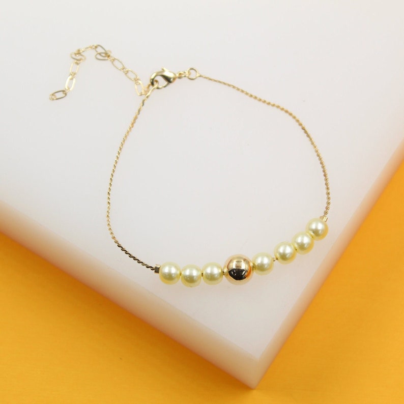 A delicate gold chain bracelet features a central arrangement of alternating pearls and a polished gold bead resting on a white display block.