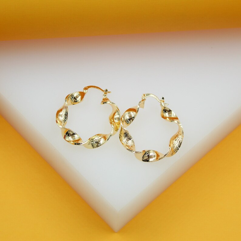 A pair of polished gold twist hoop earrings rests on a white display stand against a vibrant yellow background.
