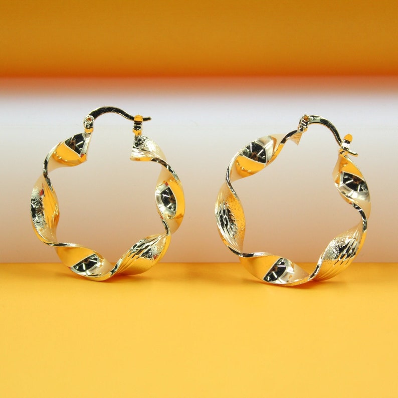 A pair of shiny gold twisted hoop earrings rests on a gradient orange and white surface.