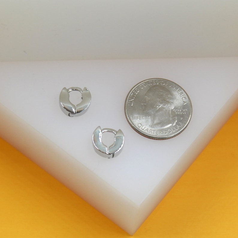 Two small silver huggie hoop earrings rest on a white surface next to a US quarter for size comparison.