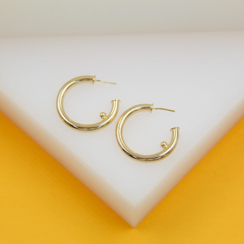 A pair of polished gold hoop earrings featuring a small spherical bead detail rests on a white display surface.