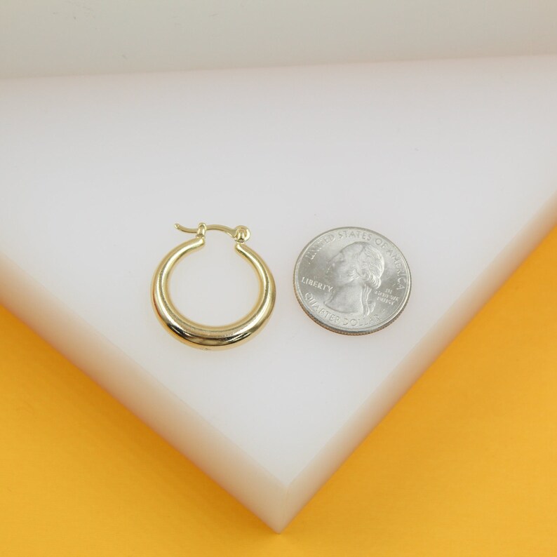 A small gold hoop earring rests on a white display next to a US quarter coin for scale.