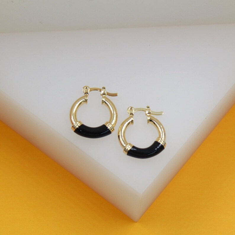 A pair of gold huggie hoop earrings featuring sleek black enamel details rests on a white geometric display.