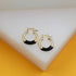 A pair of gold huggie hoop earrings featuring sleek black enamel details rests on a white geometric display.
