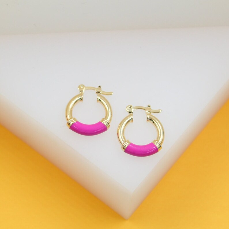 A pair of gold huggie hoop earrings featuring vibrant pink enamel details rest on a clean white and yellow display stand.