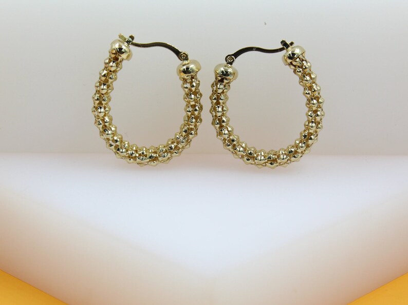 A pair of textured gold hoop earrings rests against a clean white and light orange display background.