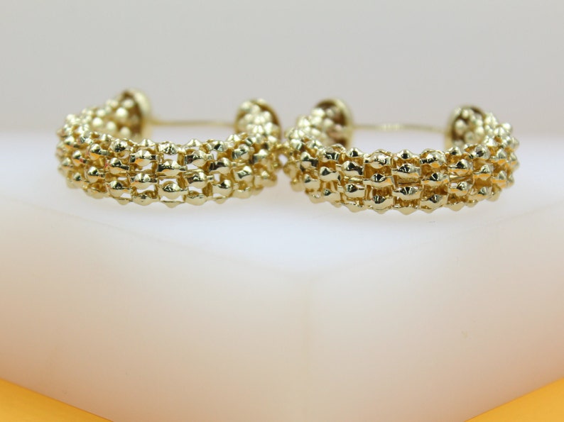 A pair of textured gold hoop earrings rests upright on a light surface, showcasing their intricate beaded design.