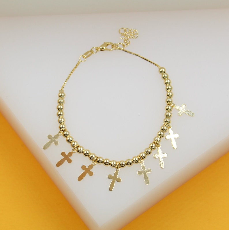 A gold beaded charm bracelet features dangling cross pendants against a neutral white background.