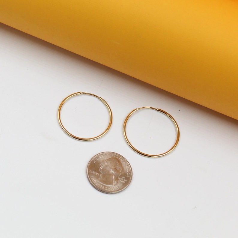 Two thin gold hoop earrings rest on a white surface next to a US quarter for size comparison.