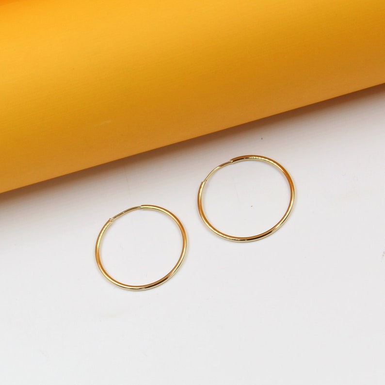 Two polished gold hoop earrings rest on a white surface against a vibrant yellow background.