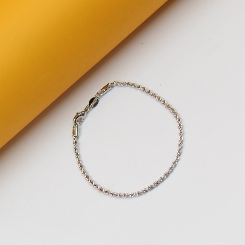 A delicate silver rope chain bracelet rests on a white and yellow surface.