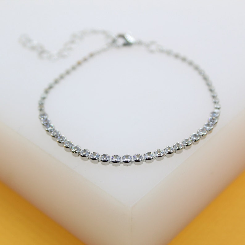 A delicate silver tennis bracelet rests on a white display stand, sparkling with a continuous line of clear cubic zirconia stones.