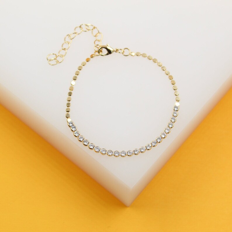 A delicate gold tennis bracelet featuring sparkling cubic zirconia stones rests on a white and yellow display block.