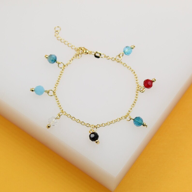 A delicate gold-link charm bracelet displays colorful multi-gemstone beads dangling against a clean white and yellow display surface.