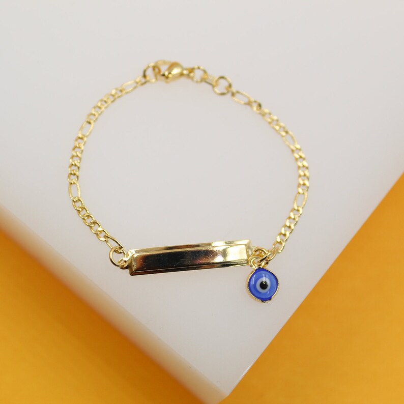 A delicate gold chain bracelet features an engraved ID bar and a dangling blue evil eye charm against a white and orange background.