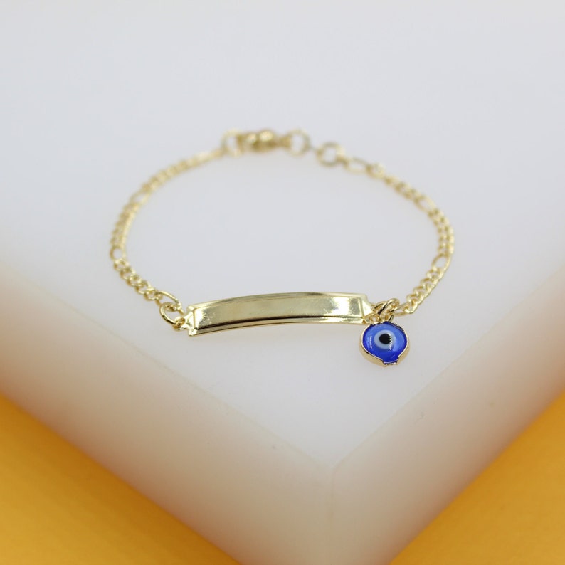 A delicate gold identification bracelet rests on a white display surface, featuring an attached blue enamel evil eye charm.