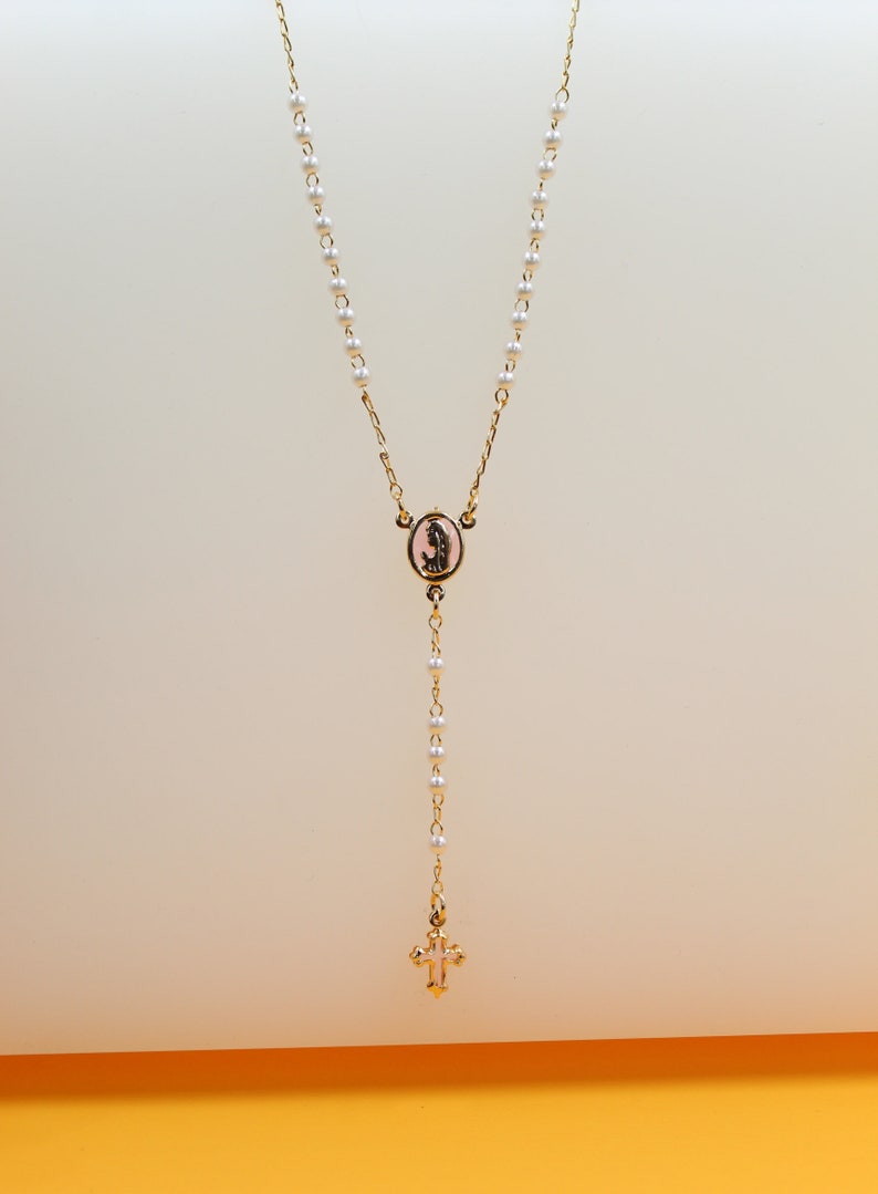 A delicate gold rosary necklace featuring small white pearls, an oval centerpiece, and a tiny cross pendant hangs against a neutral background.
