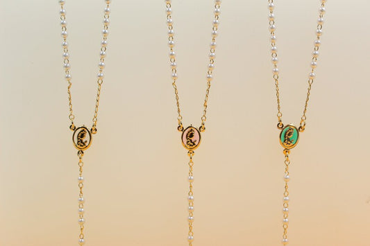 Three delicate gold-chained pearl rosary necklaces display oval centerpieces featuring a golden profile silhouette against black, pink, and green enamel backgrounds.