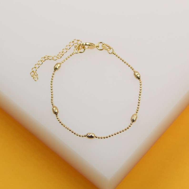 A delicate gold beaded bracelet with textured oval charms lies on a white surface.