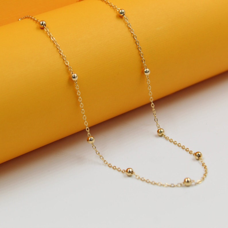 A delicate gold chain necklace with evenly spaced bead stations rests against a warm yellow and white backdrop.