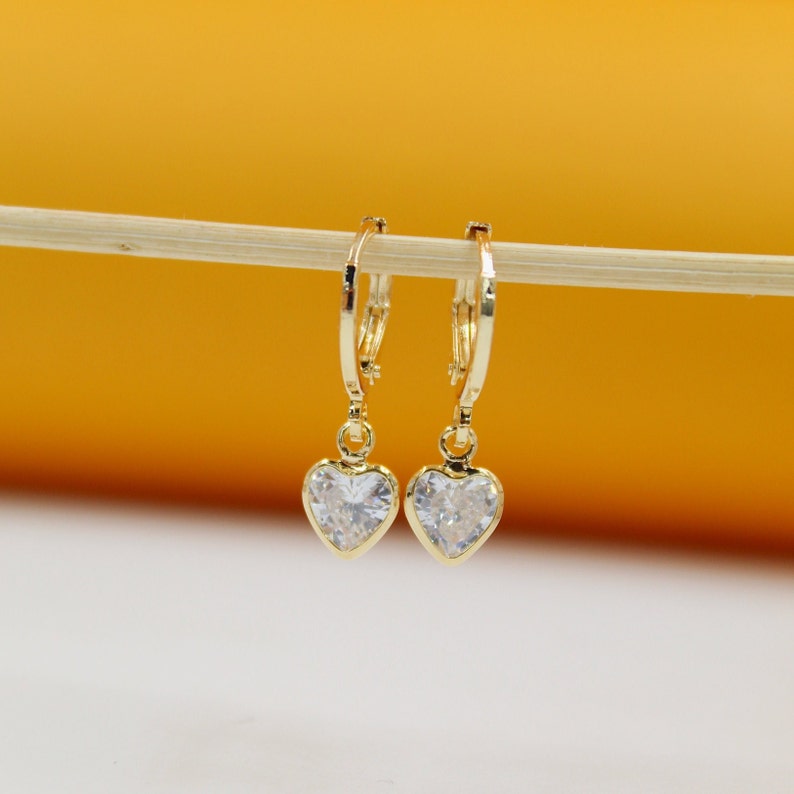 A pair of gold huggie hoop earrings featuring dangling heart-shaped crystal charms hang delicately from a wooden stick against a warm yellow and white backdrop.