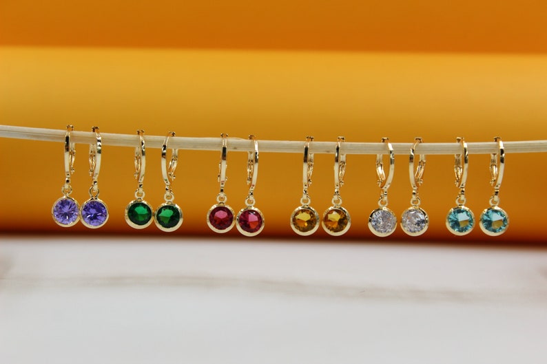 Six pairs of gold hoop earrings with colorful cubic zirconia stones hang in a row on a stick against a warm yellow and white backdrop.
