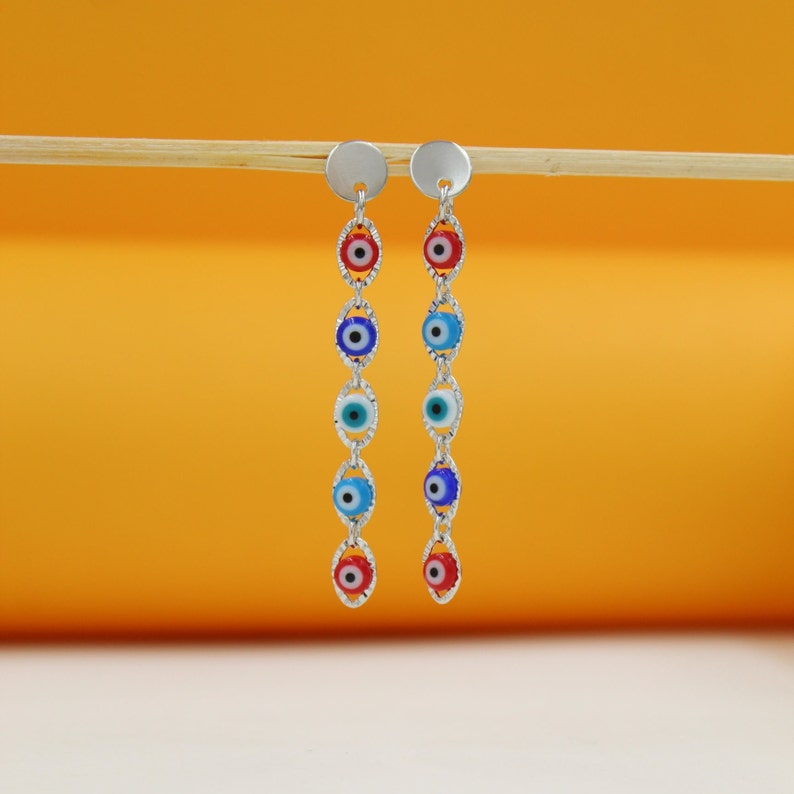 A pair of silver evil eye dangle earrings hangs from a wooden stick against a warm orange background.
