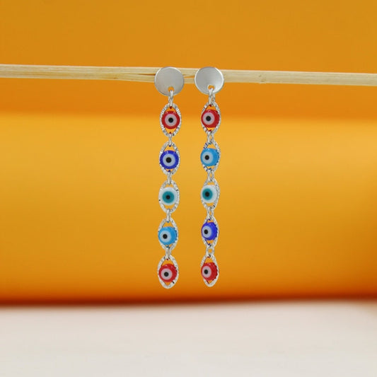 A pair of silver evil eye dangle earrings hangs from a wooden stick against a warm orange background.