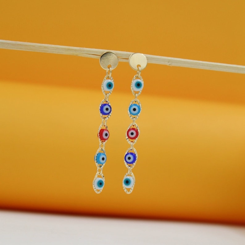 Two gold dangle earrings featuring colorful evil eye glass beads hang from a wooden stick against an orange backdrop.