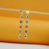 A pair of gold dangle earrings featuring multi-colored evil eye charms hangs from a thin wooden stick against a warm yellow background.