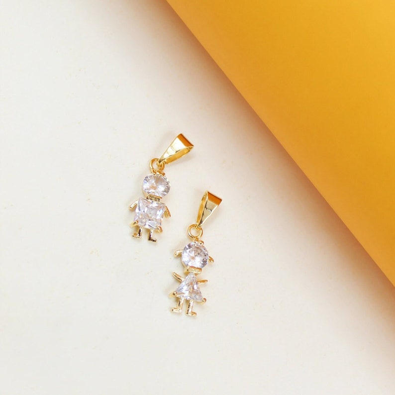 Two gold charms shaped like little girls, accented with sparkling white cubic zirconia stones, rest on a light background next to a bright yellow panel.