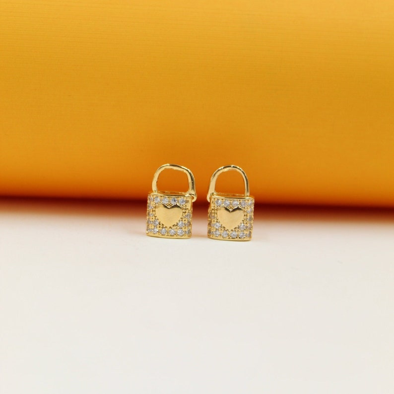 A pair of gold padlock-shaped earrings embellished with sparkling cubic zirconia crystals sits against a warm yellow and white background.