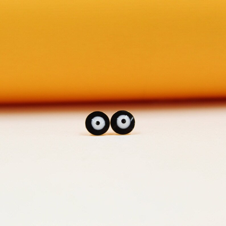 Two round black and white enamel evil eye stud earrings rest side by side against a soft peach and yellow background.