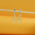 A pair of dangling gold earrings featuring intricate wire-wrapped loops hangs from a thin wooden stick against a warm orange background.