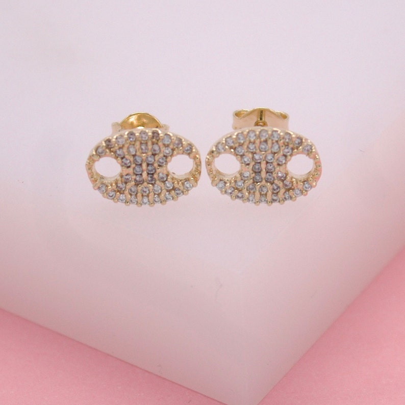 A pair of gold puff mariner link stud earrings sparkles with pave-set cubic zirconia stones against a soft white and pink background.