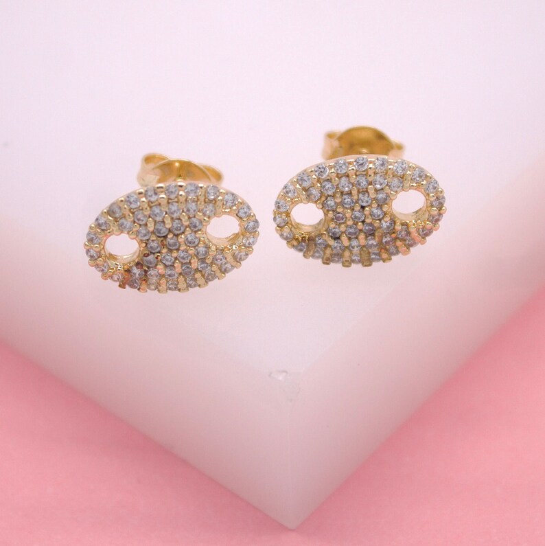 A pair of gold puff mariner link stud earrings paved with sparkling cubic zirconia rests on a white display block against a soft pink background.