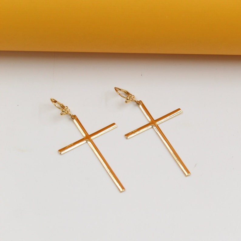 A pair of elegant gold cross earrings rests on a clean white surface against a warm yellow backdrop.