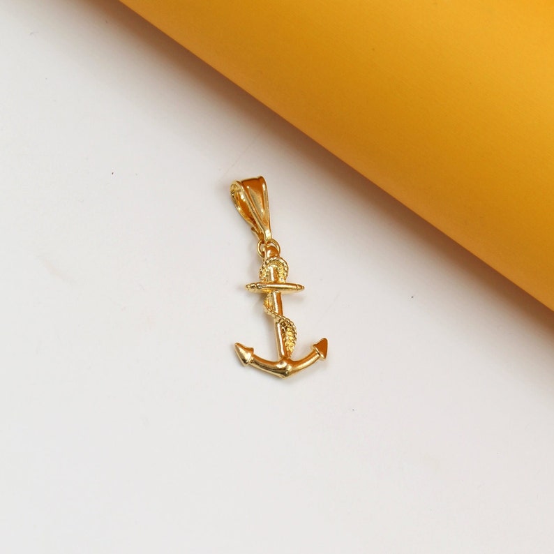 A shiny gold anchor pendant rests on a clean white surface next to a warm yellow backdrop.