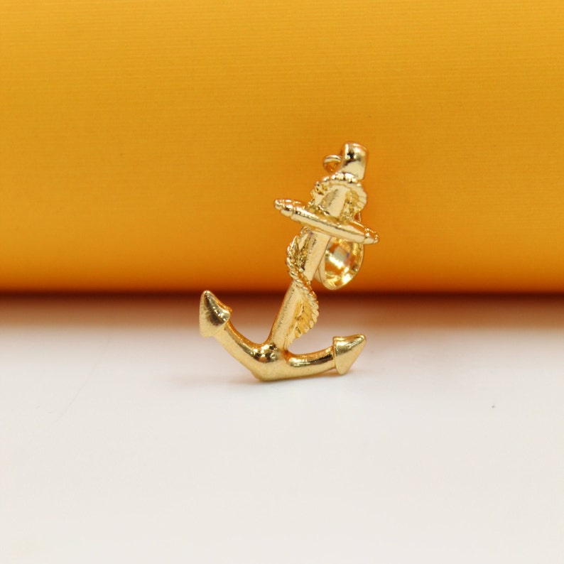 A polished gold anchor pendant stands upright against a contrasting orange and white background.