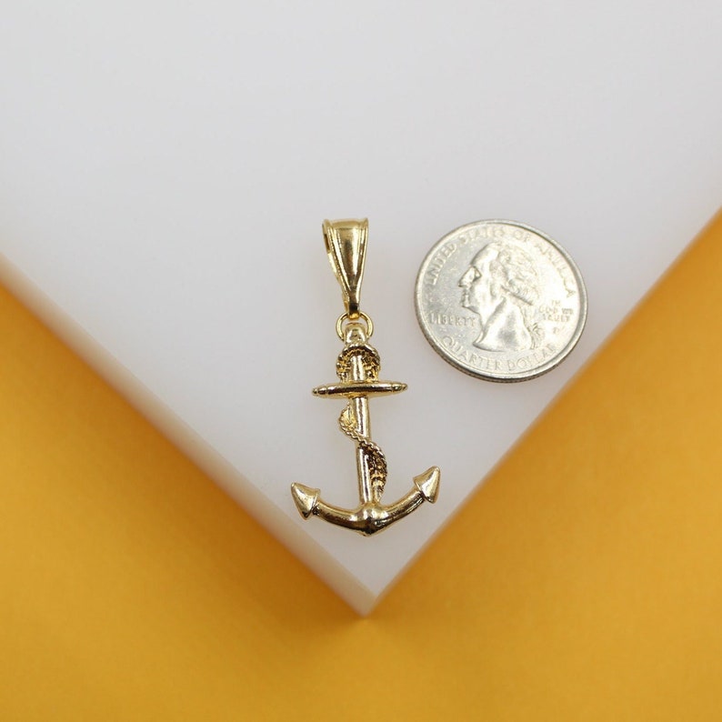A polished yellow gold anchor pendant rests beside a US quarter for scale on a white display block.